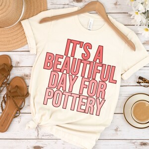 May include: A white t-shirt with the text "It's a beautiful day for pottery" in pink lettering.