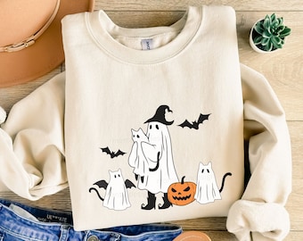 Puede incluir: Sudadera de color crema con un diseño temático de Halloween. El diseño incluye un fantasma con sombrero de bruja, sosteniendo un gato, con murciélagos y otros gatos fantasma. Una calabaza naranja también forma parte del diseño. La sudadera está sobre una superficie de madera.