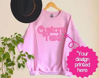 May include: A light pink sweatshirt with the words "Custom Text" in a retro font. A pink starburst graphic with the text "Your design printed here" is to the right. A black hat hangs above the sweatshirt.