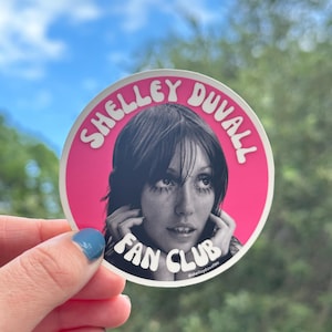Puede incluir: Una pegatina redonda con borde rosa y las palabras "SHELLEY DUVALL FAN CLUB" en blanco. La pegatina presenta una imagen en blanco y negro de Shelley Duvall. La pegatina está sujeta por una mano con esmalte de uñas azul.