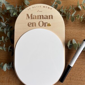 May include: A wooden memo board with a white dry erase surface. The board is shaped like a half circle and has the text "Le Petit Memo d'une Maman en Or" engraved on it. A small gold heart is also engraved on the board.