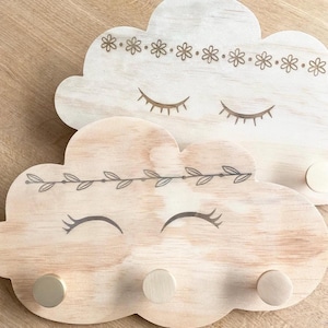 Peut inclure: Deux crochets muraux en bois en forme de nuage avec un design fantaisiste. Le crochet du haut présente une rangée de petites fleurs et des yeux fermés. Le crochet du bas a une guirlande de feuilles et des yeux fermés. Les deux crochets ont trois chevilles dorées.