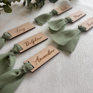Peut inclure: Cartes de place en bois avec des noms écrits en calligraphie, attachées avec un ruban vert sauge. Les noms sont : 'Emy', 'Delphine', 'Amandine', 'Marie', 'Emile' et 'Xavier'.