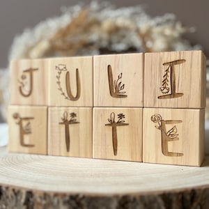 May include: Wooden blocks spelling "JULIE", each with a letter and floral etchings. The blocks are displayed on a wooden slice. The background features dried plants. The letters are decorated with botanical designs.