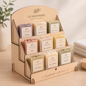 Peut inclure: Un présentoir en bois avec des savons artisanaux. Les savons sont étiquetés avec des noms comme "Lavande", "Rose" et "Patchouli". Le présentoir porte l'inscription "La Savonnerie" et "handmade with love".