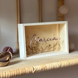 Puede incluir: Una caja de sombra de madera con el nombre "Marceau" recortado en una fuente cursiva. El interior presenta un diseño floral. La caja se exhibe en un estante tejido, con un objeto dorado en forma de corazón a la izquierda.