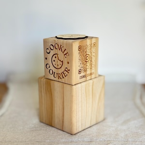 Puede incluir: Dos cubos de madera apilados con diseños grabados con láser. El cubo superior presenta las palabras "Cookie Courier" alrededor de un gráfico de galleta y un código QR con el nombre de usuario de Instagram "cookie_courier". Una pequeña galleta de madera encima.