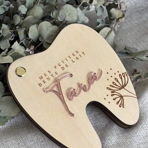 Op de afbeelding: Een houten tandvormig aandenken met de naam "Tara" in roségouden script. De tekst "MES PETITES DENTS DE LAIT" is boven de naam gegraveerd. Een kleine gouden klinknagel en een bloemmotief zijn ook zichtbaar.