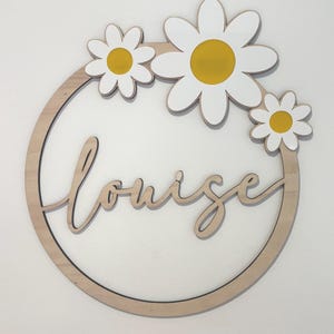 May include: A wooden circle with the name "Louise" written in cursive script. The circle is decorated with three white daisies with yellow centers.