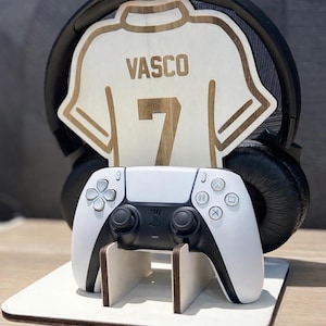 Peut inclure: Présentoir en bois avec une manette de jeu blanche et noire, un casque audio et un maillot en bois avec le nom "VASCO" et le numéro "7". La manette repose sur une base blanche.