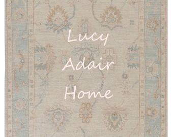 May include: A light beige rug with a blue and brown patterned design. The rug has a border with a repeating pattern. The text "Lucy Adair Home" is written in white on the rug.