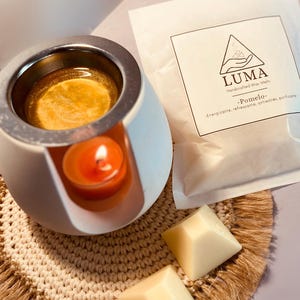 Puede incluir: Un calentador de cera de cerámica blanca con una vela de té naranja encendida en su interior. El calentador contiene cera derretida y está junto a un paquete de ceras perfumadas con el logotipo "LUMA" y el texto "Pomelo". Dos ceras cuadradas están sobre un posavasos tejido.