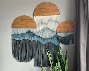 May include: A wall hanging made of wood and yarn with a wave design. The hanging is made of three circular pieces of wood with yarn fringe hanging from the bottom. The yarn is a gradient of blue and white.