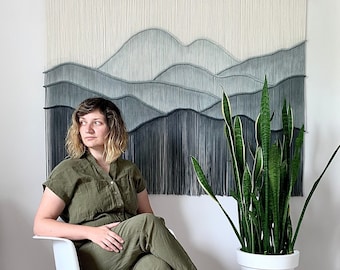 May include: A woman sits in a white chair with wooden legs, wearing a green jumpsuit. Behind her is a macrame wall hanging with a blue and white ombre design. A potted snake plant sits on a wooden plant stand to the right.