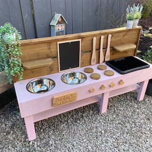 May include: A pink wooden play kitchen with a black chalkboard, two stainless steel sinks, and a black stovetop. The kitchen has a wooden shelf and a sign that reads "Sadie's Mud Kitchen".