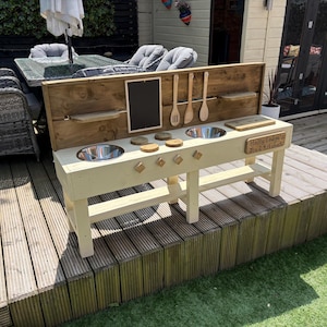 May include: A wooden outdoor play kitchen with two stainless steel sinks, wooden utensils, and a chalkboard. The play kitchen is painted in a light cream colour and has a wooden backsplash. The kitchen is designed for children.