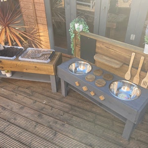 May include: A wooden outdoor play kitchen with two stainless steel sinks, a chalkboard, and a shelf for storing utensils. The kitchen is painted grey and has wooden knobs. There are two wooden planters with dirt and gravel in the background.