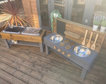 May include: A wooden outdoor play kitchen with two stainless steel sinks, a chalkboard, and a shelf for storing utensils. The kitchen is painted gray and has wooden knobs. There are two wooden planters with dirt and gravel in the background.