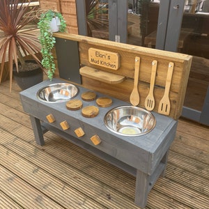 May include: A grey wooden outdoor play kitchen with two stainless steel bowls, wooden utensils, and a sign that reads "Elari's Mud Kitchen".