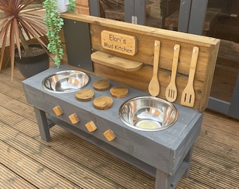 May include: A gray wooden outdoor play kitchen with two stainless steel bowls, wooden utensils, and a sign that reads "Elari's Mud Kitchen".
