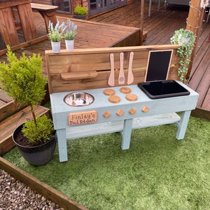 May include: A light blue wooden play kitchen with a stainless steel sink, a black plastic sink, and a wooden stovetop with four burners. The kitchen has a wooden shelf and a chalkboard. The kitchen is personalised with the name "Finlay's Mud Kitchen".
