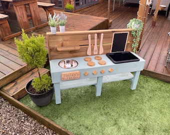 May include: A light blue wooden play kitchen with a stainless steel sink, a black plastic sink, and a wooden stovetop with four burners. The kitchen has a wooden shelf and a chalkboard. The kitchen is personalized with the name "Finlay's Mud Kitchen".
