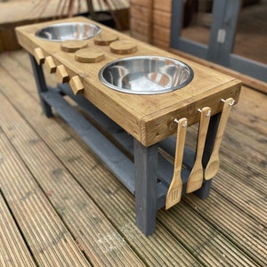 May include: Wooden play kitchen with two stainless steel sinks, four wooden utensils hanging on hooks, and a wooden countertop with round cutouts. The play kitchen is on a wooden deck.