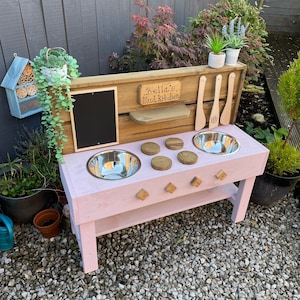 May include: A pink wooden mud kitchen playset with two stainless steel sinks, a chalkboard, and a sign that reads "Bella's Mud Kitchen".