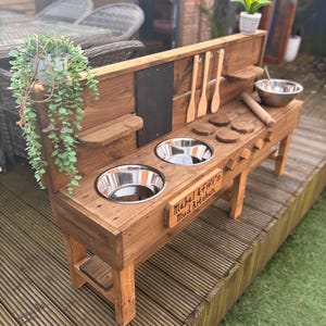 May include: A wooden outdoor play kitchen with two stainless steel bowls, a chalkboard, and a sign that reads "Mabel & Fifi's Mud Kitchen".