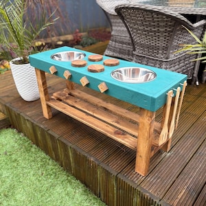 May include: A teal blue wooden play kitchen with two stainless steel sinks, wooden knobs, and a shelf. The kitchen is on a wooden deck and has wooden utensils hanging on the side.