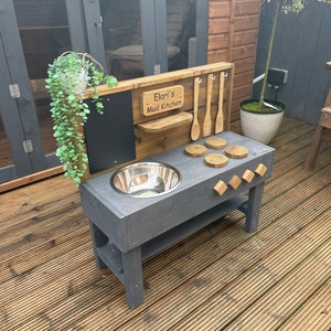 May include: A grey wooden outdoor play kitchen with a stainless steel sink, a chalkboard, and a sign that reads "Elani's Mud Kitchen".