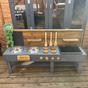 May include: A wooden outdoor play kitchen with a black painted finish. It has two stainless steel bowls, a black plastic sink, and a wooden sign that reads "Elari's Mud Kitchen".