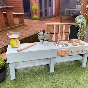 May include: A light blue wooden play kitchen with a sink, a rolling pin, and a sign that says "Tiney's Mud Kitchen".