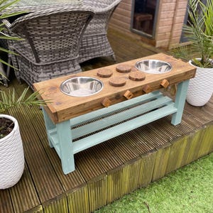 May include: A wooden pet feeding station with two stainless steel bowls. The top is stained wood, and the base is painted a pale blue. Includes four wooden coasters and six wooden pegs. The unit is on a wooden deck with potted plants.