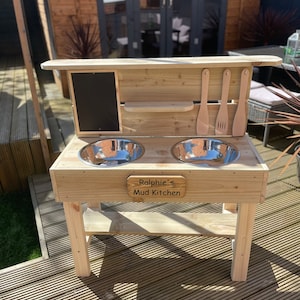 May include: Wooden mud kitchen playset with two stainless steel bowls, a chalkboard, and a shelf. The kitchen has a sign that reads "Ralphie's Mud Kitchen".