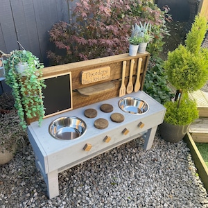 May include: A grey wooden mud kitchen with stainless steel bowls and wooden utensils. The kitchen has a chalkboard, wooden burners, and the text "Theodore Mud Kitchen". The outdoor play set is surrounded by greenery and sits on a bed of gravel.