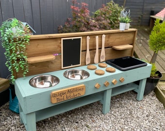 May include: A light blue wooden mud kitchen playset with two stainless steel bowls, a black chalkboard, and wooden spoons hanging on hooks. The kitchen has a sign that reads "Unique Abilities - Mud Kitchen".