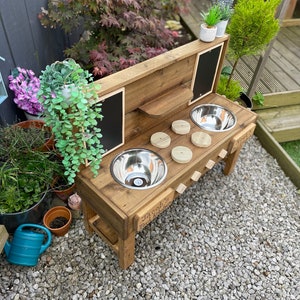 May include: A wooden play kitchen with two stainless steel sinks, a shelf, and two chalkboard panels. The kitchen is personalised with the name "Jacob's Mud Kitchen".