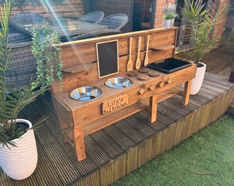 May include: A wooden outdoor play kitchen with two stainless steel sinks, a chalkboard, and wooden utensils. The kitchen is labeled "Lily's Mud Kitchen".