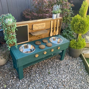 May include: A green wooden mud kitchen with a chalkboard, a sign that says "Theodore Mud Kitchen", two stainless steel bowls, and wooden knobs. The kitchen is set up outdoors on a gravel surface.