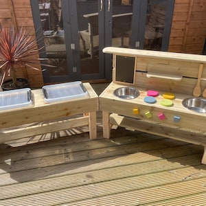 May include: Wooden outdoor play kitchen with two stainless steel sinks, a chalkboard, and two trays. The kitchen is painted in natural wood tones and has a shelf for storage.