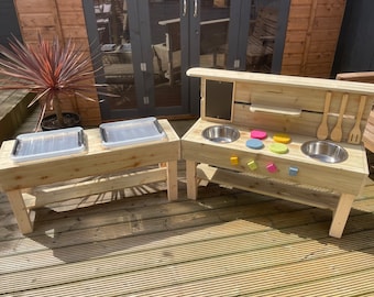 May include: Wooden outdoor play kitchen with two stainless steel sinks, a chalkboard, and two trays. The kitchen is painted in natural wood tones and has a shelf for storage.