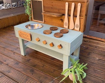 May include: A light blue wooden play kitchen with a stainless steel sink, a chalkboard, and wooden utensils hanging on the side. The kitchen has a sign that says "Rosie's Mud Kitchen".