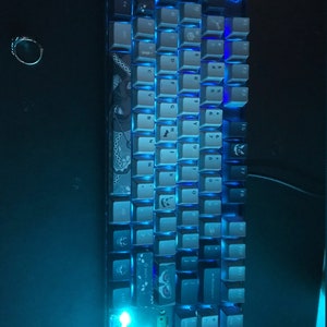 May include: A mechanical keyboard with blue LED backlighting. The keycaps are a mix of gray and white, with some keys featuring decorative designs. A red keycap is visible in the top right corner. The keyboard is designed for gaming or computer use.