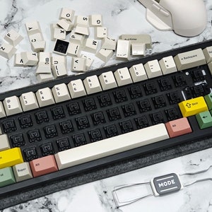 May include: A black mechanical keyboard with off-white keycaps and colorful accent keys. The keyboard has a variety of keys, including yellow, pink, and green. A keycap puller and a white mouse are also present.