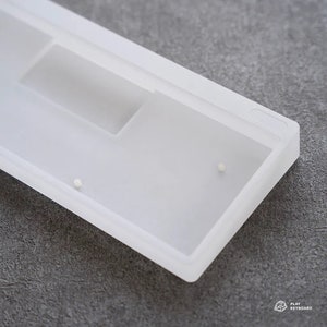Puede incluir: Una carcasa de teclado blanca translúcida con un diseño rectangular. La carcasa tiene un acabado liso y mate y presenta recortes para componentes. Se ven dos pequeñas clavijas blancas en la superficie. El texto "PLAY KEYBOARD" es visible.
