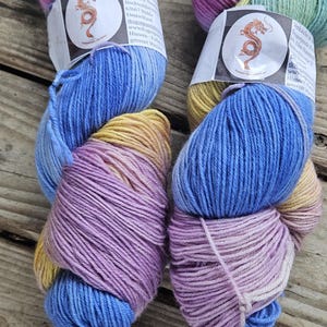 May include: Two skeins of hand-dyed yarn featuring a variegated colour palette of pink, blue, yellow, and purple. Each skein has a white paper label with a circular dragon logo and text including the website dragonpanda.de and washing instructions.