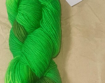 May include: A skein of vibrant green yarn with varying shades, from bright to deep green. A tag with text is attached. The yarn is tightly wound, showcasing its texture and color variations. Ideal for knitting or crochet projects.