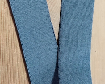 Pode incluir: Uma banda elástica de cor cinza escura, dobrada ao meio, com uma superfície texturizada. A banda tem aproximadamente 2,5 cm de largura.