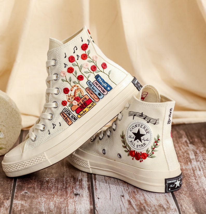 May include: A pair of white high-top Converse sneakers with embroidered floral designs and music notes. One shoe features a design of books with song titles and artist names, including "folklore", "Lover", "EVERMORE", "Midnighk", "49.89", "RE", "Taylor Swift", "SPEAK NOW", and "1989". The other shoe features a Converse All Star logo with a floral design.